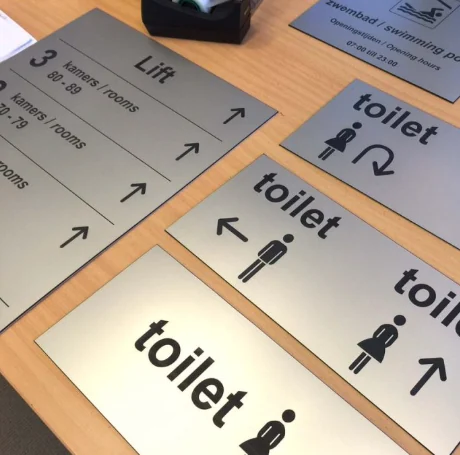 Gegraveerde borden met pijlen en aanduidingen voor kamers, lift en toiletten.