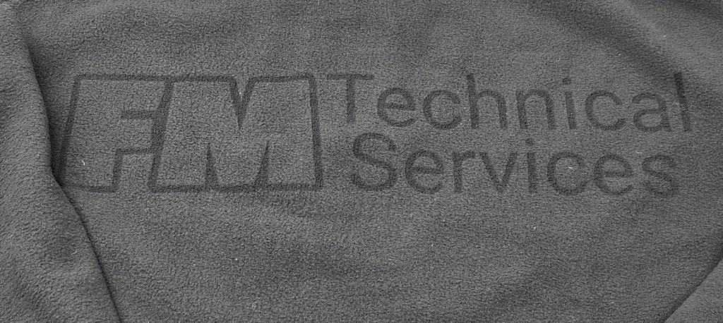 Close-up of the dark FM Technical Services logo in the texture of a fleece jacket.